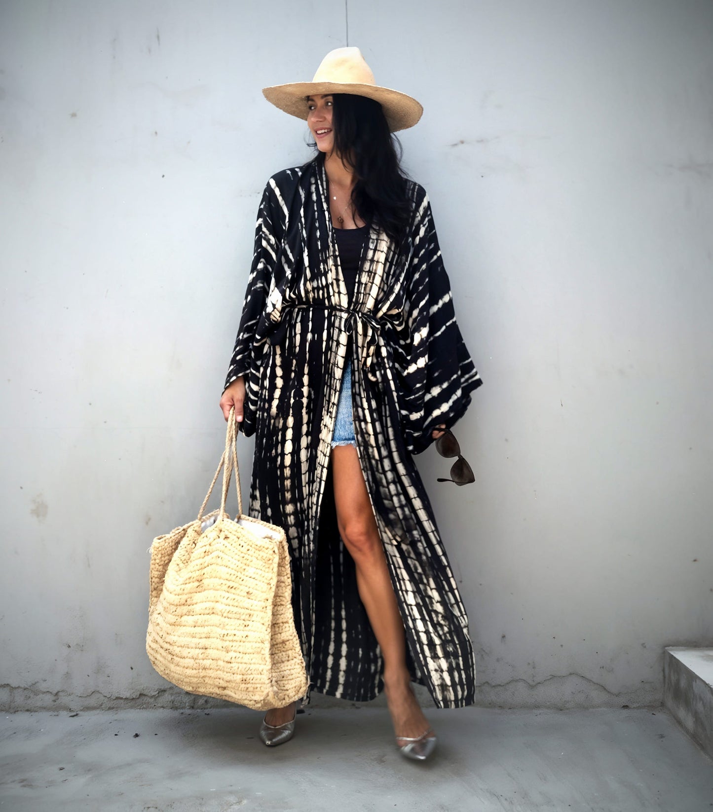 Woman wearing a long black and white patterned coat, holding a straw bag, against a plain wall.
