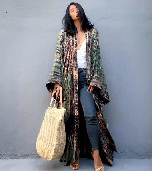 Woman wearing a long, patterned kimono over a white top and blue jeans, holding a straw bag against a gray background.