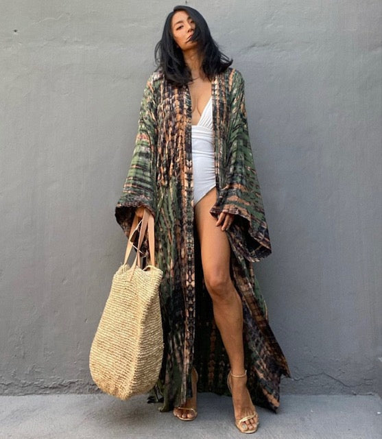 Woman wearing a patterned kimono over a white outfit, holding a straw bag against a gray wall.