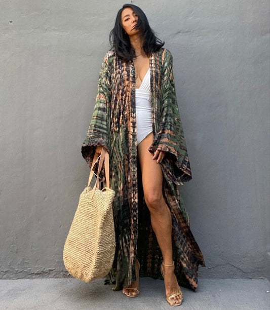 Woman wearing a patterned kimono over a white outfit, holding a straw bag against a gray wall.